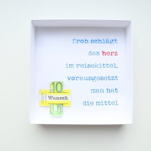 May include: A white frame with a white background and blue and red text that reads "froh schl&auml;gt das herz im reisekittel, vorausgesetzt man hat die mittel". A yellow sticker with the word "Wunsch" and the number "10" is attached to the frame.