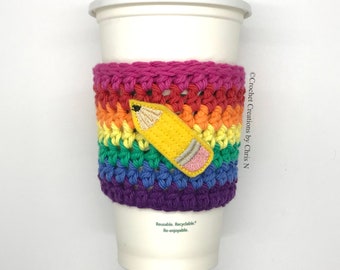 May include: A reusable coffee cup with a rainbow-colored, crocheted cozy. The cozy features a yellow pencil applique. The cup is white with a white lid. The text "Reusable. Recyclable. Re-enjoyable." is printed on the cup.