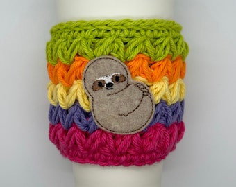 May include: A colorful, handmade crochet coffee cup sleeve. The sleeve features a rainbow pattern with green, orange, yellow, purple, and pink stripes. A felt sloth applique is centered on the sleeve. The sleeve is designed to fit a standard coffee cup.