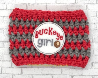 May include: A handmade crochet coffee cup cozy in red and gray. The cozy features a white circular patch with the words "Buckeye Girl" embroidered in red and gray, with a brown buckeye graphic. The cozy is designed to fit around a standard coffee cup.