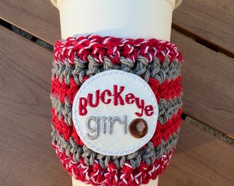 May include: A white reusable coffee cup featuring a crocheted sleeve with alternating horizontal stripes of red and grey yarn. A circular white patch is attached to the center, embroidered with the red and grey text Buckeye girl.