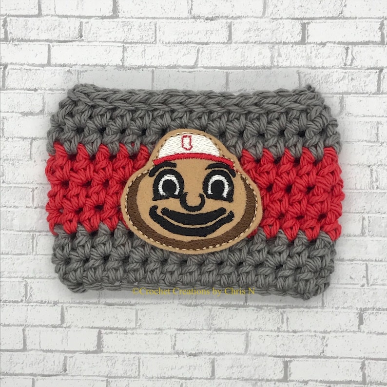 May include: A handmade gray and red crocheted coffee cup cozy featuring a brown and tan Ohio State Buckeyes mascot applique. The cozy is rectangular with a red stripe in the middle and a mascot in the center.