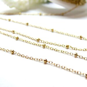 Peut inclure: Gros plan de plusieurs colliers délicats de couleur or. Chaque collier est composé d'une fine chaîne entrecoupée de petites perles dorées espacées régulièrement. Les chaînes sont disposées en diagonale sur un fond blanc, créant une sensation de profondeur et de texture.