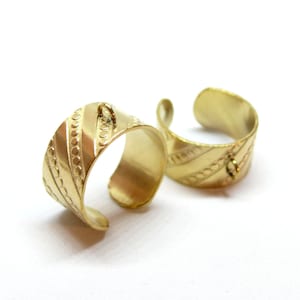 Op de afbeelding: Twee goudkleurige verstelbare ringen. Elke ring heeft een brede band met geëtste diagonale lijnen en kleine cirkelvormige patronen. Aan de bovenkant van elke ring is een kleine lus bevestigd. De ringen worden op een witte achtergrond weergegeven.