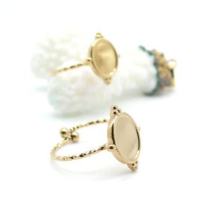 Peut inclure: Bagues réglables dorées avec un sertissage ovale. Les bagues ont un anneau texturé et de petits détails décoratifs autour de l'ovale. Les bagues sont présentées sur fond blanc.