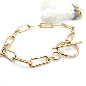 Op de afbeelding: Goudkleurige schakelarmband met een tuimelsluiting. De armband heeft langwerpige rechthoekige schakels en een cirkelvormige sluiting met een rechte staafsluiting. De sieraden worden op een witte achtergrond gepresenteerd.