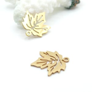 Könnte beinhalten: Zwei goldfarbene Blatt-Anhänger mit einem Ausschnitt-Design. Die Anhänger haben oben eine kleine Schlaufe zum Befestigen an Schmuck. Die Blätter sind mit Adern detailliert und haben ein mattes Finish.