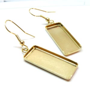 Può includere: Una coppia di basi per orecchini in metallo color oro con castoni rettangolari dotati di base piatta e incassata. Ogni orecchino include una chiusura a gancio semplice fissata alla parte superiore della cornice rettangolare da un anellino.