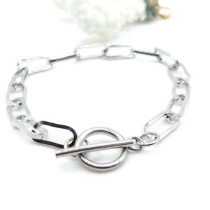 May include: A silver-toned chain bracelet featuring elongated rectangular links and a circular toggle clasp closure. The bracelet is constructed from polished metal links, with one link appearing in a darker, matte black finish, creating a contrast.