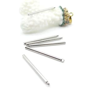 Op de afbeelding: Een verzameling zilverkleurige metalen sieradenonderdelen, waaronder verschillende rechte, cilindrische staven met afgeronde uiteinden. Eén staaf heeft een kleine lus aan het uiteinde. De onderdelen zijn verspreid op een wit oppervlak, met een wit, getextureerd object op de achtergrond.