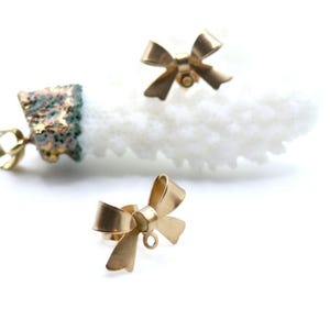 Può includere: Orecchini dorati a forma di fiocco. Gli orecchini sono piccoli e hanno una finitura lucida. I fiocchi sono disposti su uno sfondo bianco, con un oggetto simile al corallo sullo sfondo. Gli orecchini sono probabilmente per orecchie forate.