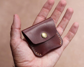 May include: A small, dark brown leather pouch with a brass button closure. The edges are stitched with yellow thread. The pouch is held in an open hand, showcasing its compact size and craftsmanship. A minimalist design for carrying small items.