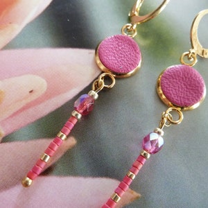 Puede incluir: Un par de pendientes de cuero rosa con aros dorados y cuentas de cristal rosa. Los pendientes cuelgan y tienen un círculo de cuero rosa en la parte inferior.