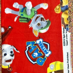 May include: A red fleece fabric featuring a repeating Paw Patrol pattern with characters like Rocky, Marshall, and Rubble, alongside the blue and white Paw Patrol logo. The white selvage edge includes the text 2015 Spin Master Used under license.