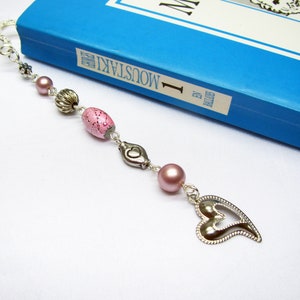 Könnte beinhalten: Ein silbernes Lesezeichen mit einem Herz-Anhänger, rosa und silbernen Perlen und einer silbernen Kette. Das Lesezeichen hängt an einem blauen Buch mit dem Titel "Moustaki I" auf dem Rücken.