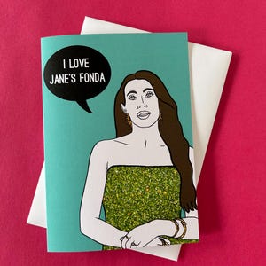 Può includere: Biglietto di auguri con sfondo turchese, con l'illustrazione di una donna in un abito verde glitterato. Un fumetto nero recita "I LOVE JANE'S FONDA". Il biglietto è abbinato a una busta bianca, su sfondo rosa.