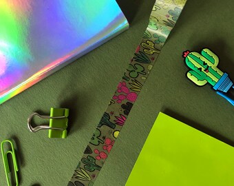 Op de afbeelding: Een groene en bruine washi tape met een cactuspatroon. De tape ligt op een groene ondergrond met een holografisch notitieboek, groene paperclips en een groen en geel cactusfiguurtje.