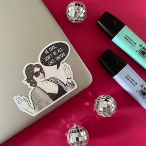 Könnte beinhalten: Ein holografischer Aufkleber zeigt eine Person mit Sonnenbrille und dunklem Oberteil sowie eine Sprechblase mit dem Text, BE COOL, DON'T BE ALL, LIKE, UNCOOL. Der Aufkleber klebt auf einem silbernen Laptop neben drei kleinen Discokugeln und zwei Textmarkern.