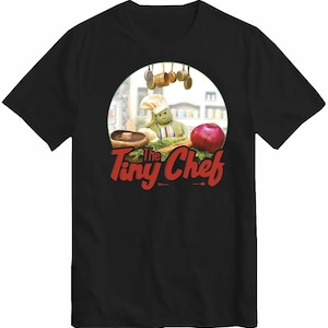 May include: Black t-shirt with a circular graphic of a cartoon chef. The chef, wearing a white hat and apron, is surrounded by vegetables. The text "The Tiny Chef" is in red script.
