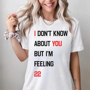 May include: White t-shirt with the text "I DON'T KNOW ABOUT YOU BUT I'M FEELING 22" in black and red. The person in the image is wearing a gold necklace and a scrunchie on their wrist.