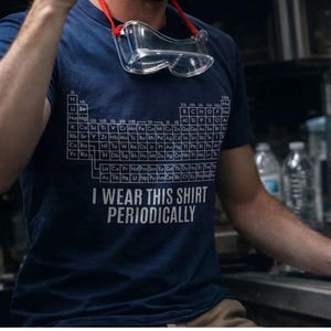 May include: A navy blue t-shirt featuring a white graphic of the periodic table of elements and the text "I WEAR THIS SHIRT PERIODICALLY" printed in white. Clear safety goggles with a red strap are resting around the neck of the shirt.