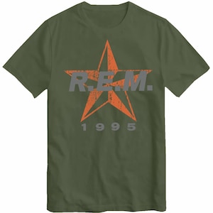 May include: An olive green t-shirt with a distressed orange star design. The graphic includes the letters "R.E.M." in grey and the number "1995". The shirt has a crew neck and short sleeves.