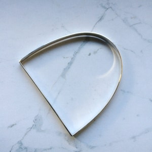 May include: A silver-coloured metal cookie cutter in a distinctive teardrop shape. The cutter features a rounded top and straight sides, ideal for baking. The cutter is presented on a white marble surface.