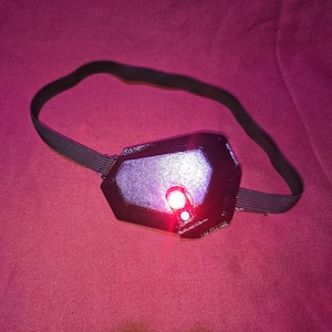 May include: A black, futuristic-looking eye patch with a red light and a black elastic band. The eye patch has a geometric design and appears to be made of plastic or a similar material. The red light is illuminated.