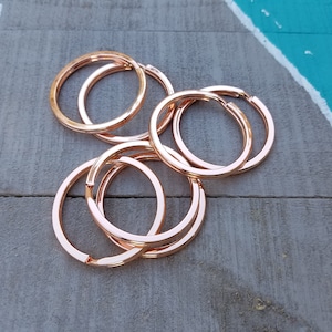 May include: A group of six rose gold key rings, stacked and overlapping. The key rings are made of metal and have a smooth, polished finish.