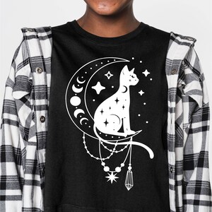May include: A black cropped t-shirt featuring a white graphic of a cat sitting on a crescent moon with stars and a dangling crystal charm.