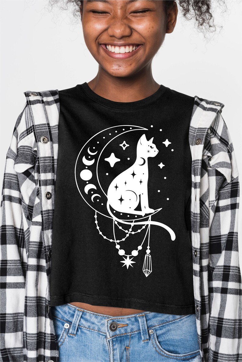 May include: A black cropped t-shirt featuring a white graphic of a cat sitting on a crescent moon with stars and a dangling crystal charm.