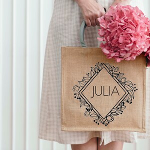 May include: A brown burlap tote bag with a black floral design and the name "JULIA" printed in a diamond shape. The bag is being held by a person wearing a white dress.