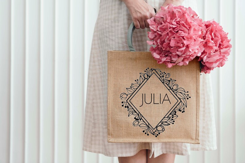 May include: A brown burlap tote bag with a black floral design and the name "JULIA" printed in a diamond shape. The bag is being held by a person wearing a white dress.