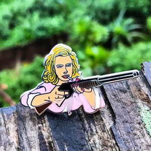 May include: A pink enamel pin featuring a woman with blonde hair holding a shotgun and smoking a cigarette. The pin is set against a wooden background.
