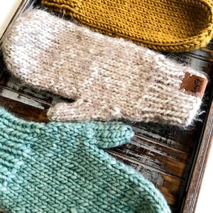 Op de afbeelding: Drie paar gebreide wanten in een houten dienblad. De wanten zijn in verschillende kleuren en texturen: een lichtbruin paar, een lichtbeige paar met witte vlekken en een tealgroen paar. Elk paar heeft een klein leren label met het woord "Cozy" erop.