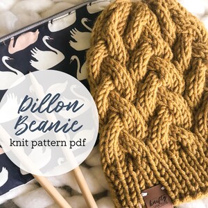 May include: A close-up of a mustard yellow knitted beanie with a cable knit pattern. The beanie is lying on a white surface with a black and white fabric with white swans printed on it. The text "Pillon Beanie knit pattern pdf" is written in white on a white circle. Two wooden knitting needles are visible in the image.