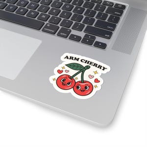 May include: A white sticker featuring two smiling red cherries with green stems and a leaf. The sticker has the text "ARM CHERRY" above the cherries, with hearts and stars around the image. The sticker is on a laptop.