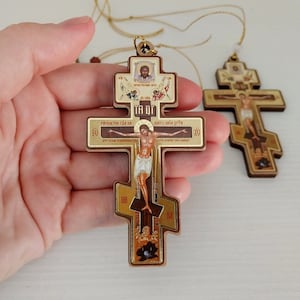 May include: Two wooden Orthodox crosses with gold detailing and religious imagery. The main cross depicts Jesus Christ, with gold inscriptions. The crosses are suspended by gold cords. The crosses are approximately 10 cm in length.