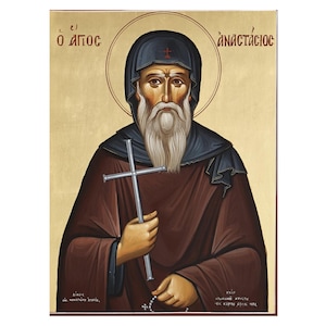 May include: An icon of a religious figure with a long white beard, wearing a dark brown robe and a black head covering. The figure holds a silver cross and is set against a gold background. Greek text is visible at the top.