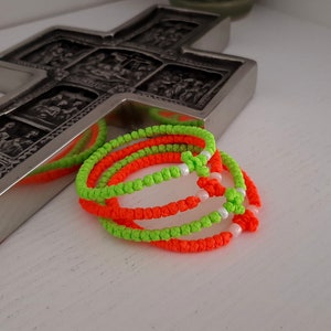 Può includere: Una croce d'argento con immagini religiose dettagliate sullo sfondo. In primo piano, diverse corde di preghiera arancioni e verde neon brillante, ciascuna con perline bianche. Le corde sono annodate e sembrano fatte a mano.