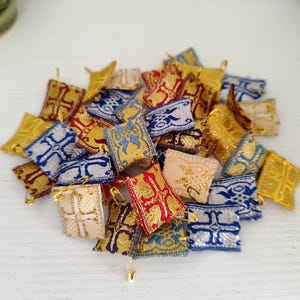 Könnte beinhalten: Eine Ansammlung kleiner, dekorativer Stoffquadrate in Gold, Rot, Blau und Creme. Jedes Quadrat weist aufwendige Muster auf und ist mit einer Sicherheitsnadel versehen. Die Quadrate sind in einem Haufen angeordnet, was auf ein Bastel- oder Verzierungsobjekt hindeutet.