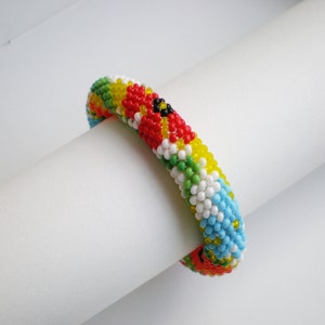 May include: A colorful beaded bracelet with a pattern of red, yellow, green, blue, and white beads. The bracelet is made of small, round beads and is woven in a tight pattern.