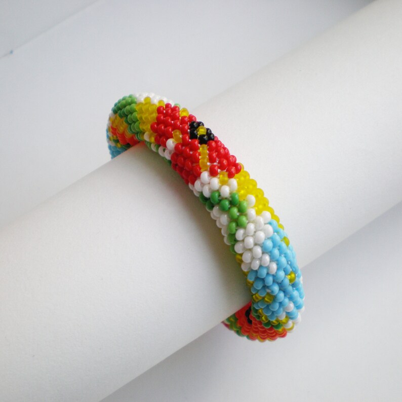 May include: A colorful beaded bracelet with a pattern of red, yellow, green, blue, and white beads. The bracelet is made of small, round beads and is woven in a tight pattern.