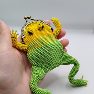 May include: A beaded frog-shaped coin purse in yellow and green. The frog has a yellow head and green body, with a metal clasp. The purse is held in a hand, showcasing the detailed beadwork and whimsical design.