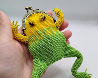 May include: A beaded frog-shaped coin purse in yellow and green. The frog has a yellow head and green body, with a metal clasp. The purse is held in a hand, showcasing the detailed beadwork and whimsical design.