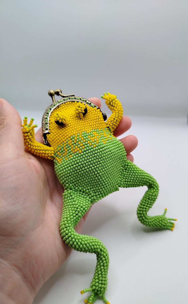 May include: A beaded frog-shaped coin purse in yellow and green. The frog has a yellow head and green body, with a metal clasp. The purse is held in a hand, showcasing the detailed beadwork and whimsical design.