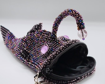 May include: A beaded pouch shaped like an anglerfish, covered in iridescent dark purple and multi-colored beads. It features a curved lure with a clear bead at the tip, two light purple circular eyes, and a black zippered mouth with a metal keyring.