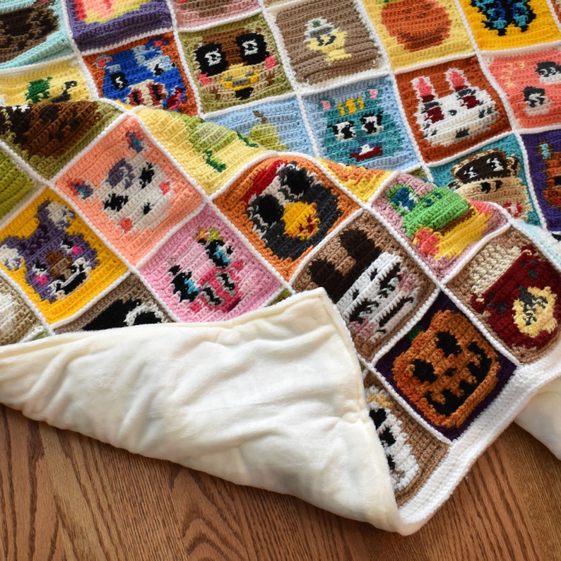 May include: A crocheted blanket with a variety of colorful squares featuring animal characters. The blanket is folded in half, revealing a white fleece lining.