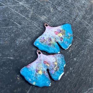 Könnte beinhalten: Zwei handgefertigte, blau-weiße Emaille-Blattanhänger mit bunten floralen Akzenten. Jeder Anhänger hat oben eine kleine Schlaufe zum Aufhängen. Die Anhänger werden auf einer dunklen, strukturierten Oberfläche präsentiert.