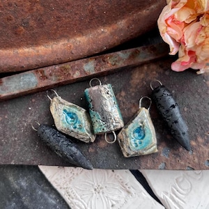 May include: Five ceramic pendants with metal loops for hanging. Two are long, textured black cones. Two are diamond-shaped with a light blue glazed centre. One is a rectangular cylinder with a metallic blue and brown finish. A pale pink rose is nearby.